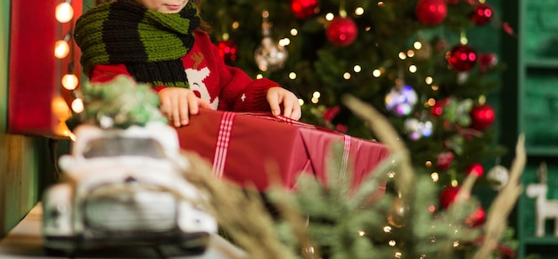 Un ragazzino con un maglione rosso e una sciarpa verde si siede con un regalo di Natale tra le mani nell'arredamento serale di Natale. Foto