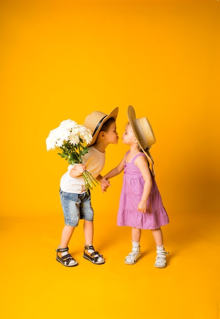 Un ragazzino con un mazzo di fiori bacia una bambina con un cappello di paglia su una superficie gialla con spazio per il testo