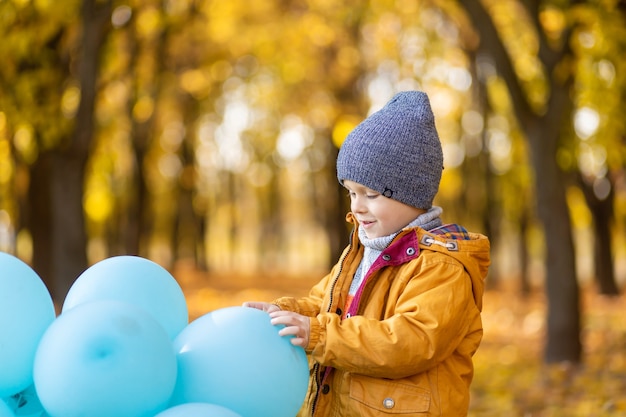 Un ragazzino con una bracciata di palloncini cammina nel parco autunnale. Alberi gialli e palline blu. Bambino alla moda.