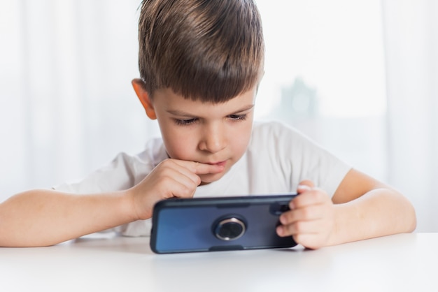 Un ragazzino con una maglietta bianca gioca al telefono a casa. Un bambino felice guarda il suo smartphone.