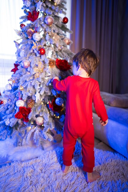 Un ragazzino con una tuta da neve rossa si trova di fronte a un albero di Natale L'albero è decorato con ornamenti rossi e dorati e il ragazzo sta tendendo la mano per toccarlo