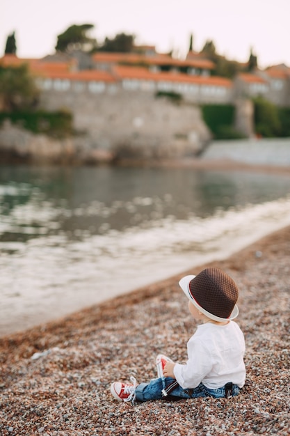 Un ragazzino dagli occhi azzurri con un cappello carino è seduto in riva al mare sullo sfondo dell'isola di Sveti Stefan, vista posteriore.