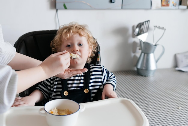 Un ragazzino dai capelli ricci si siede su una sedia da bambino in cucina, la madre nutre un porridge sano