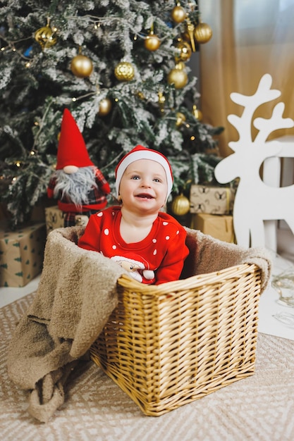 Un ragazzino di 1 anno seduto in un cestino sullo sfondo di un albero di Natale Un bambino nel vento rosso di Capodanno vicino all'albero di Natale