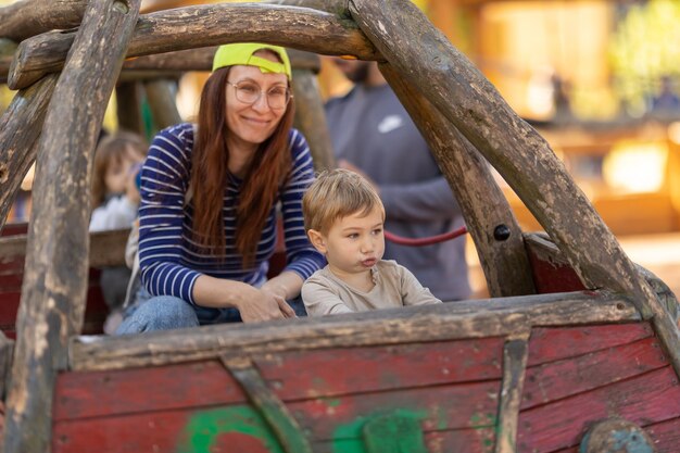 Un ragazzino divertente e sua madre sorridente seduti nella barca di legno nel parco giochi