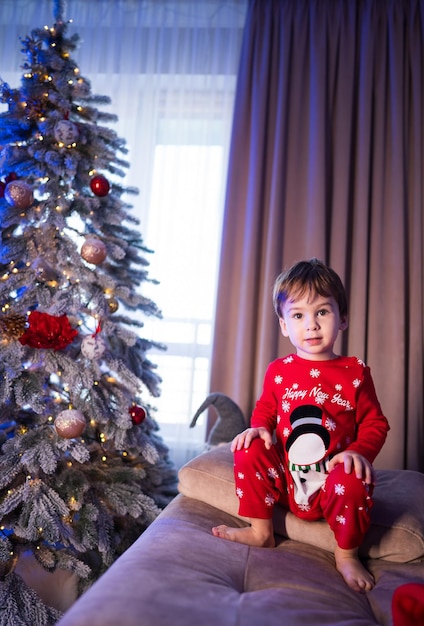 Un ragazzino è seduto su un divano davanti a un albero di Natale indossa una tuta da neve rossa e si sta godendo l'atmosfera festiva