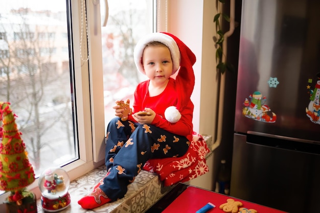 Un ragazzino ha indossato un cappello da Babbo Natale ed è seduto a casa vicino alla finestra con dei dolci