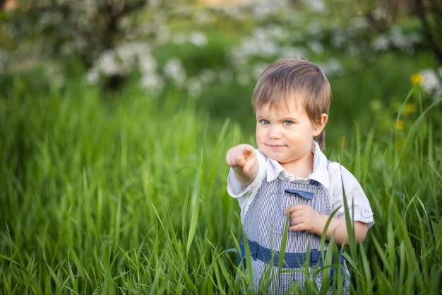 Un ragazzino in salopette di jeans con gli occhi azzurri espressivi. Saltare e scherzare nell'erba alta e verde sullo sfondo di un grande cespuglio verde e di un giardino fiorito.