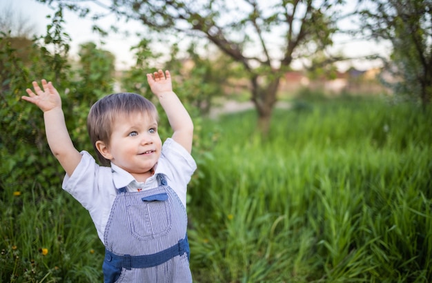 Un ragazzino in salopette di jeans con gli occhi azzurri espressivi. Saltare e scherzare nell'erba alta e verde sullo sfondo di un grande cespuglio verde e di un giardino fiorito.