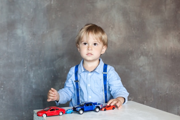 Un ragazzino in una camicia con bretelle gioca con macchinine giocattolo colorate. Ragazzo prescolare che gioca con l'automobile del giocattolo su una tavola a casa o sull'asilo nido. Giocattoli educativi per bambini in età prescolare e all'asilo.