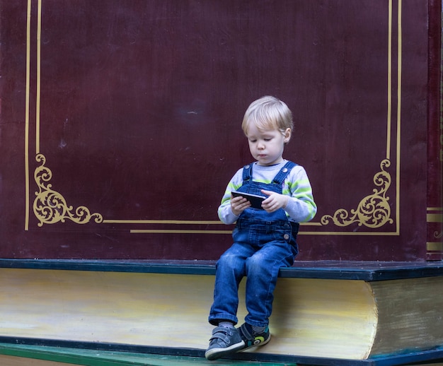 Un ragazzino si siede su un libro e guarda e gioca sullo schermo dello smartphone