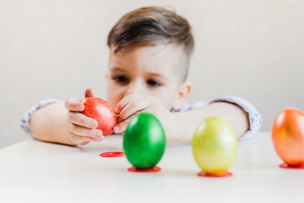 Un ragazzino su un tavolo bianco tiene tra le mani e sbuccia un uovo di Pasqua rosso su sfondo bianco.