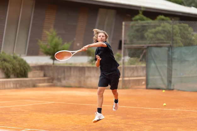 Un ragazzo adolescente in maglietta nera che gioca a tennis sul telaio del campo in movimento colpendo la palla