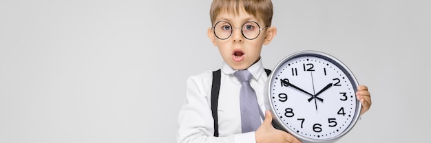 Un ragazzo affascinante con camicia bianca, bretelle, cravatta e jeans chiari è grigio. Il ragazzo tiene in mano un orologio