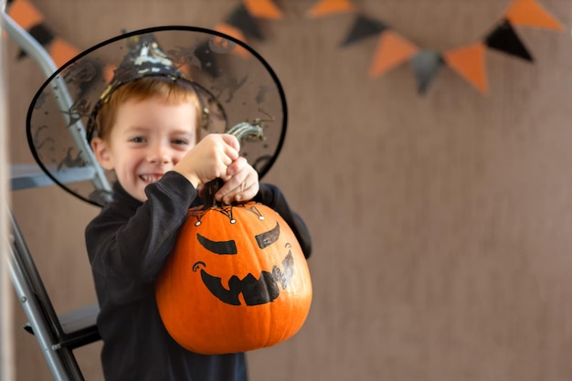 UN RAGAZZO ALLEGRO E SVEGLIO DI 5 ANNI CHE TIENE UNA ZUCCA ARANCIO CON UNA Smorfia DIPINTA PER HALLOWEEN, ALL'INTERNO CON COPYSPACE.