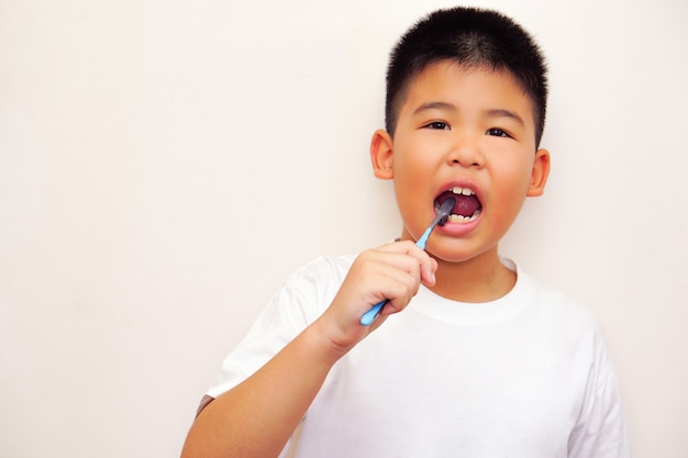 Un ragazzo asiatico con una maglietta bianca si pulisce i denti e guarda la telecamera (sfondo bianco). Concetto di igiene e pulizia.