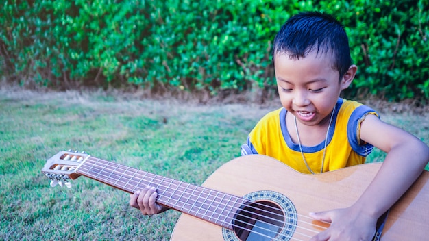 Un ragazzo asiatico sta suonando una chitarra acustica in un giardino con alberi verdi.