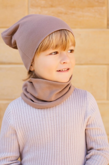 un ragazzo biondo con le fossette sulle guance con un cappello beige e uno snood sullo sfondo di un muro di pietra