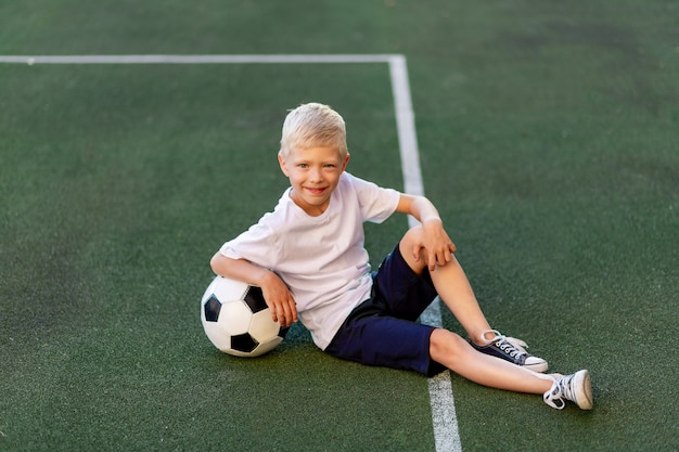 un ragazzo biondo in uniforme sportiva si siede su un campo di calcio con un pallone da calcio
