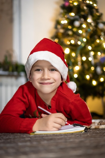 Un ragazzo carino con un maglione rosso e un cappello rosso da Babbo Natale scrive una lettera a Babbo Natale e pensa ai regali