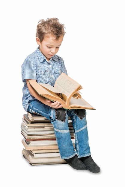 Un ragazzo carino con una camicia blu seduto su una pila di libri e che legge un libro su uno sfondo bianco isolato