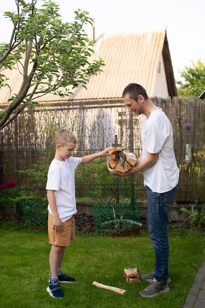 Un ragazzo carino con una maglietta bianca raccoglie legna da ardere e la dà a suo padre Legna da ardere sull'erba