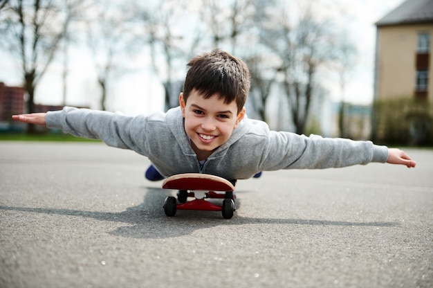 Un ragazzo carino sdraiato con la pancia su uno skateboard con le braccia tese come ali e immaginando un volo su un aeroplano