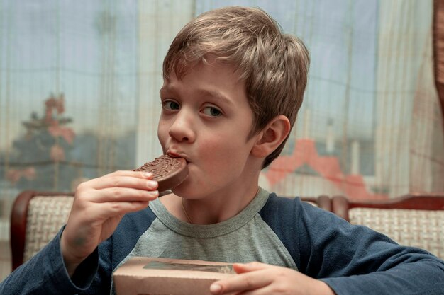 Un ragazzo caucasico allegro e carino che mangia cioccolato seduto a un tavolo in una cucina di casa con la bocca imbrattata di cioccolato