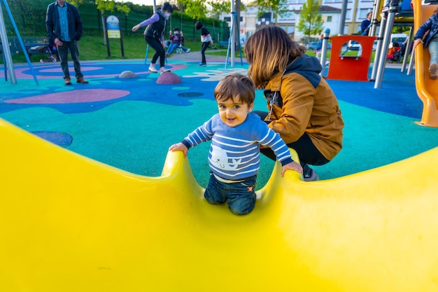 Un ragazzo caucasico di un anno che gioca ad altalene con sua madre che gioca nel parco saltando su uno squeaker giallo