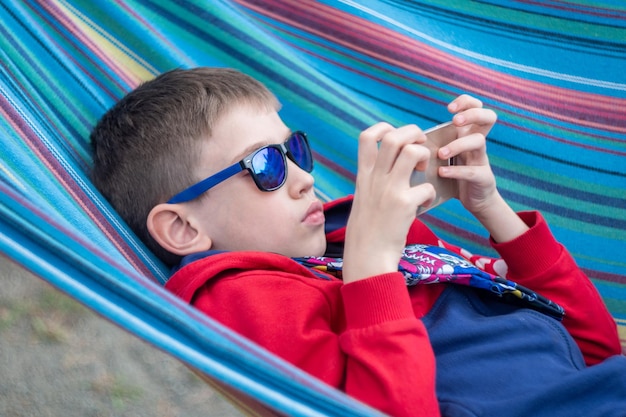 Un ragazzo con gli occhiali da sole sdraiato su un'amaca colorata e che gioca con il telefono Giochi educativi nell'app per smartphone Ora legale Il bambino usa sempre il telefono giochi di comunicazione online