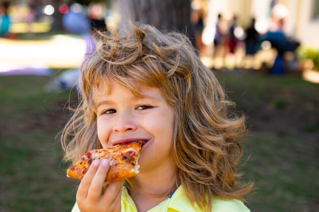 Un ragazzo con prende un boccone di pizza. Il bambino mangia una fetta di pizza ai peperoni. Fronte divertente dei bambini.