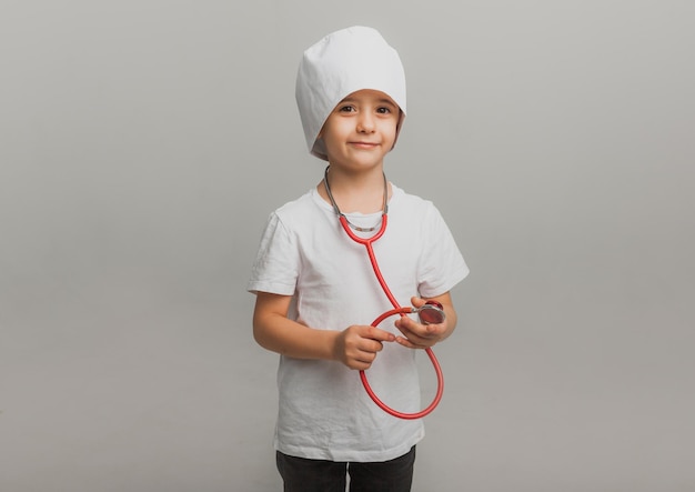 Un ragazzo con un berretto da dottore tiene uno stetoscopio su uno sfondo chiaro. piccolo dottore. foto in studio di alta qualità