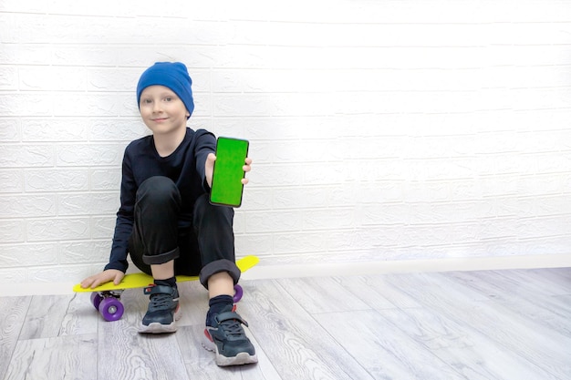 Un ragazzo con un cappello blu si siede su uno skateboard che tiene in mano un telefono con uno schermo verde