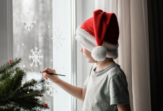 Un ragazzo con un cappello da Babbo Natale disegna fiocchi di neve sulla finestra. Tradizioni familiari natalizie