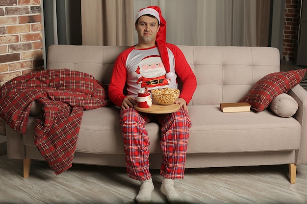 un ragazzo con un cappello da Babbo Natale e un pigiama è seduto sul divano con in mano un vassoio con popcorn