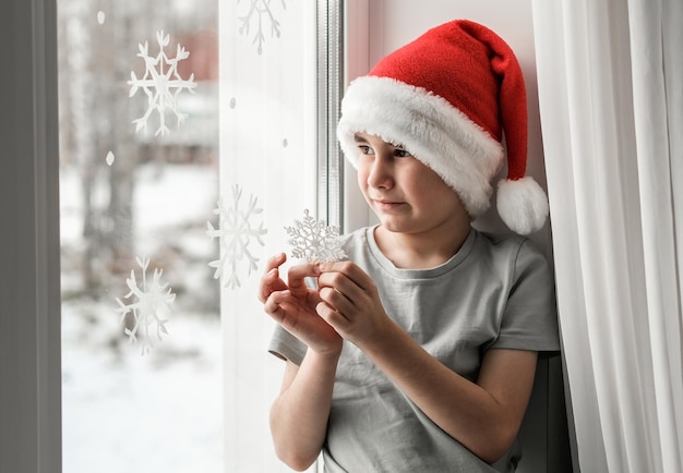 Un ragazzo con un cappello da Babbo Natale tiene in mano un fiocco di neve e guarda fuori dalla finestra