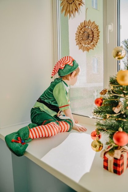 Un ragazzo con un costume da elfo di Natale dell'aiutante di Babbo Natale guarda fuori dalla finestra