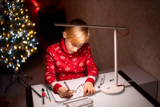 Un ragazzo con un maglione rosso di Natale sullo sfondo di un albero di Natale dipinge un disegno con pennarelli