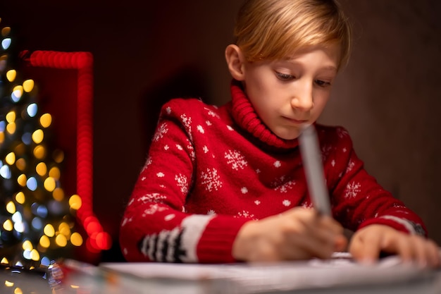 Un ragazzo con un maglione rosso di Natale sullo sfondo di un albero di Natale tiene in mano un pennarello e disegna su carta