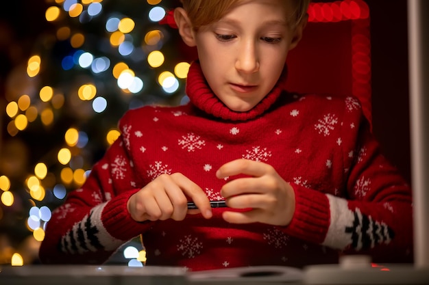 Un ragazzo con un maglione rosso di Natale sullo sfondo di un albero di Natale tiene un pennarello per disegnare