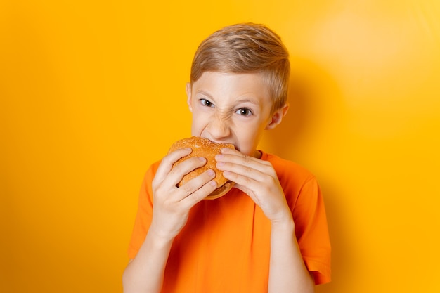 Un ragazzo con una maglietta arancione tiene un hamburger con entrambe le mani e lo morde avidamente su uno sfondo giallo