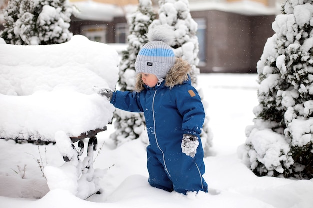 Un ragazzo con una tuta blu e un cappello vede la neve per la prima volta e si trova vicino a una panchina sullo sfondo di un albero di Natale coperto di neve nel parco.