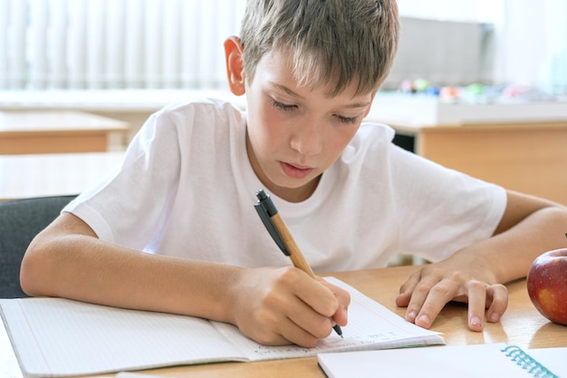 Un ragazzo concentrato che fa i compiti scrivendo il testo in un taccuino al tavolo Ragazzo carino e positivo scrive e sorride Torna al concetto di scuola Istruzione scolastica e prescolare