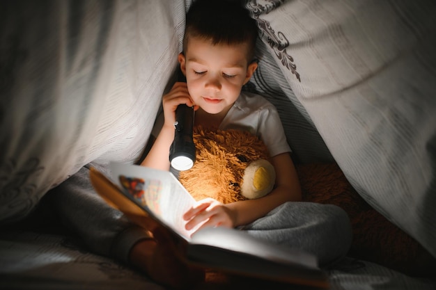 Un ragazzo di 56 anni sta leggendo un libro la sera al buio sotto una coperta con un orso giocattolo