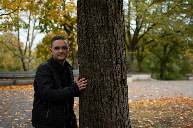 Un ragazzo giovane adulto sta nel parco guardando al lato pensieroso, in abiti scuri, nel parco verde