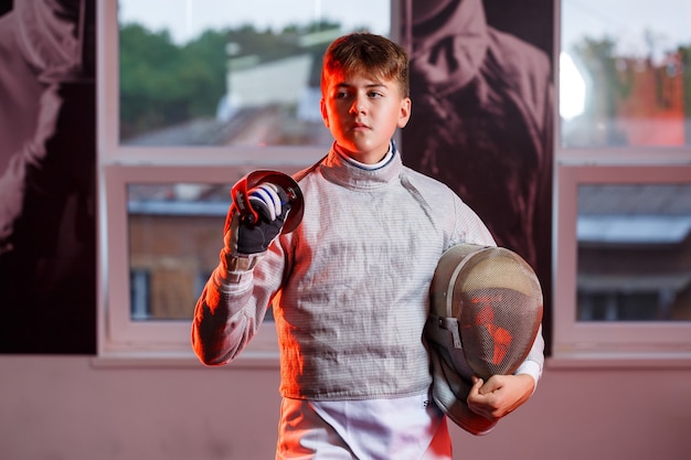 Un ragazzo in tuta da scherma con una spada in mano, luce al neon. Un giovane modello si allena e si allena in movimento, azione. Sport, gioventù, stile di vita sano.