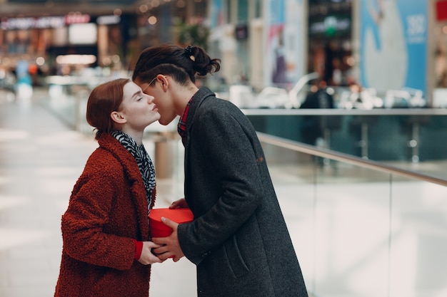 Un ragazzo regala alla sua ragazza una scatola a forma di cuore nel giorno di San Valentino. Giovani coppie che baciano al centro commerciale.