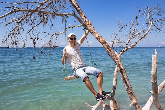 Un ragazzo seduto su un'altalena, su un albero caduto sulla riva e navi da carico all'orizzonte. La periferia della località di Gelendzhik. Russia, costa del Mar Nero