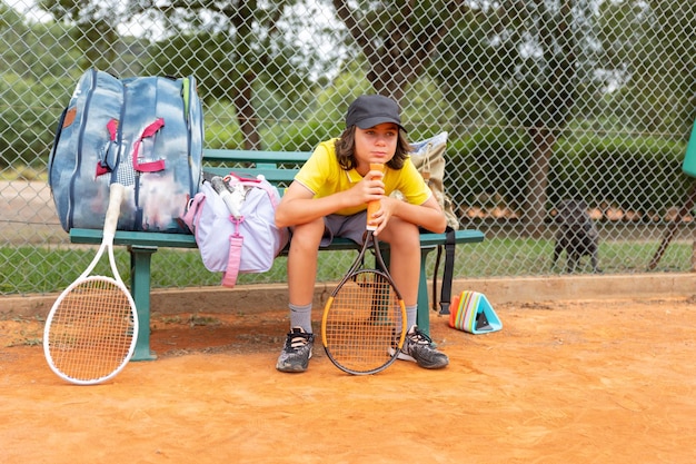 Un ragazzo seduto su una panchina con le mazze sportive vicino al campo da tennis e in attesa del suo turno
