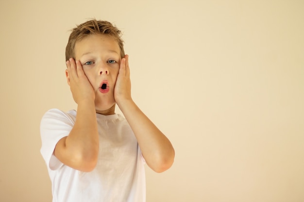 Un ragazzo sorpreso o spaventato di 7-10 anni con una maglietta bianca si alza e grida con le mani sulle guance su uno sfondo beige. Copia spazio.